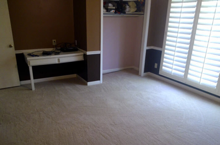 Spacious room with beige carpet, a white desk, and brown-painted walls with natural light from shuttered windows.