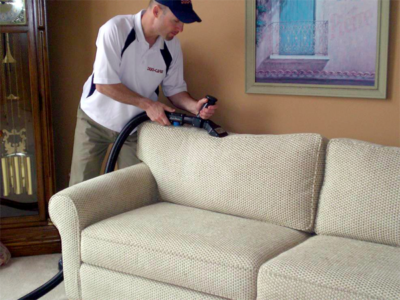 A professional cleaner using a vacuum to clean upholstery on a beige sofa in a living room.
